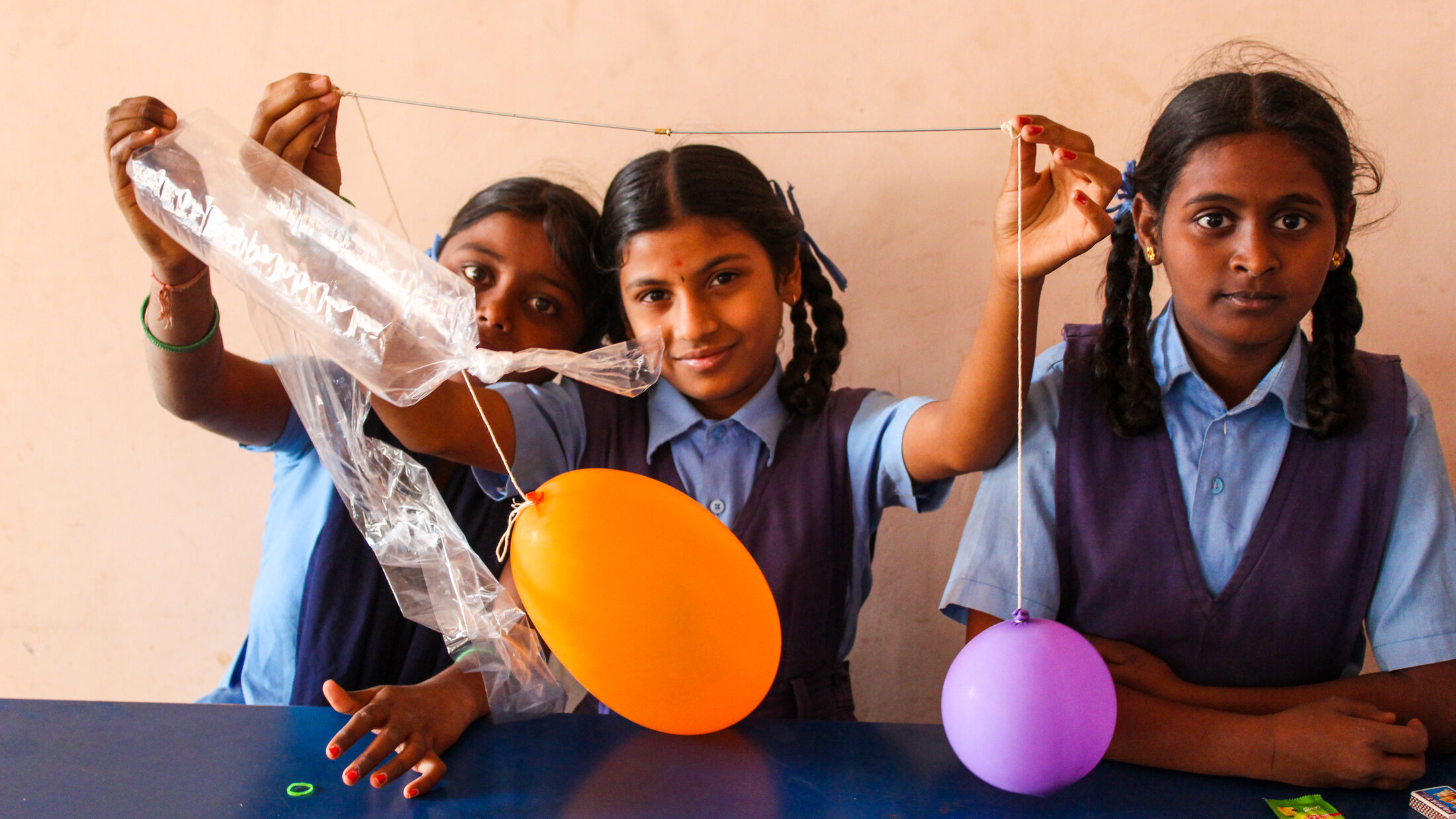 A smiling girl in school uniform holding colourful balloons during a balloon-and-bottle air-pressure experiment.